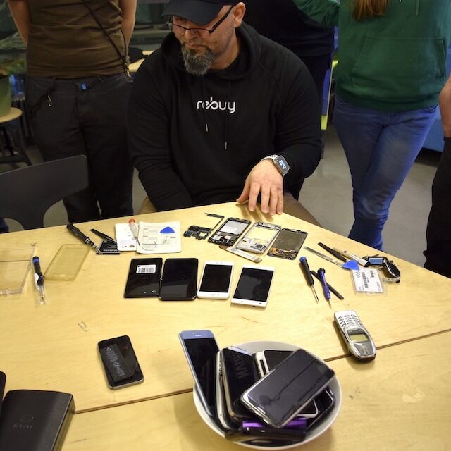 Ein Mann sitzt an einem Tisch. Vor ihm liegen verschiedene Handyteile und Werkzeuge.